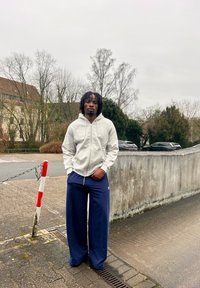 Jeune homme portant un sweat à capuche gris zippé et un pantalon large bleu marine, debout sur une rue pavée près d'arbres dénudés et de voitures garées par une journée nuageuse.