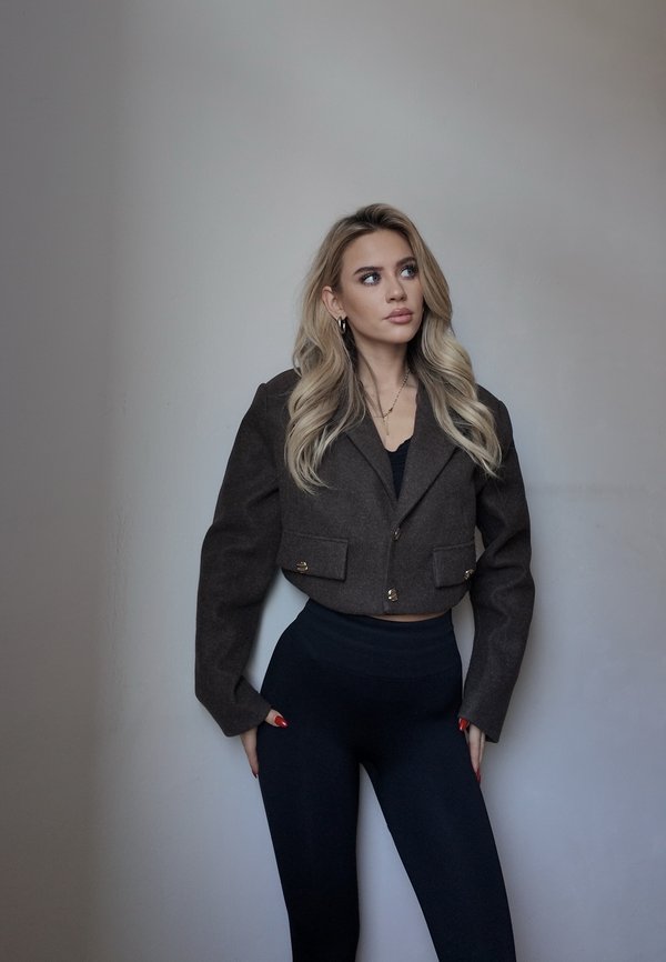 Una mujer rubia posa frente a una pared gris, con una chaqueta corta oscura y pantalones negros, mirando hacia un lado.