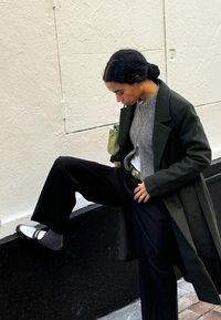 A model is wearing a long dark green coat over a grey sweater and black wide-leg trousers, paired with silver shoes and a green bag.