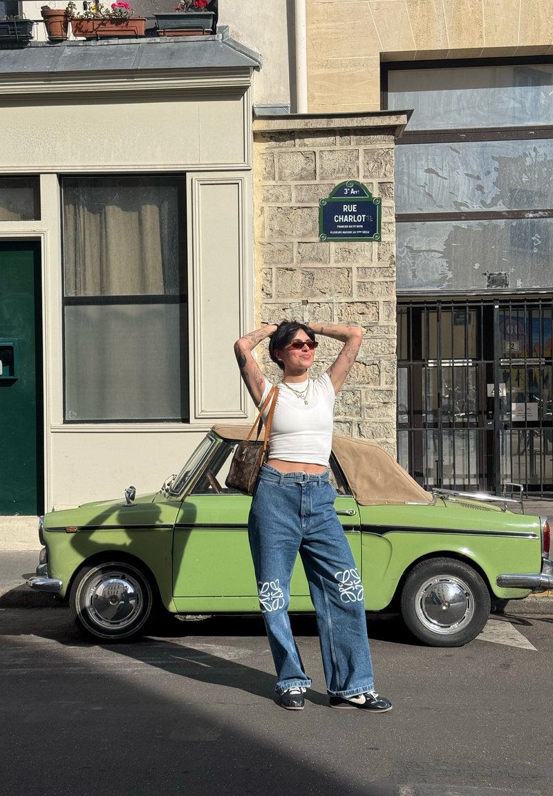 Femme en crop top blanc et jean large debout, les mains derrière la tête, à côté d'une voiture décapotable verte vintage dans la rue Rue Charlot.