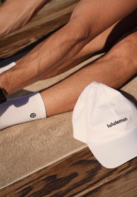 Biała czapka baseballowa Lululemon z haftowanym logo, umieszczona na beżowym ręczniku. Widoczne nogi w białych skarpetach z logo.