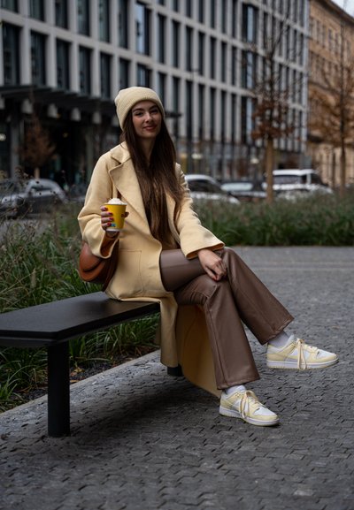 Abrigo beige largo, pantalones marrones y zapatillas amarillas con detalles en blanco. Sentado en un banco negro, sosteniendo una taza de café amarilla al aire libre.