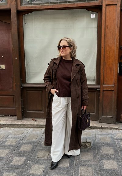 Abrigo de ante marrón sobre un suéter burdeos, combinado con pantalones anchos blancos y tacones puntiagudos negros. Bolso negro con detalles dorados.