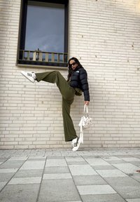 Μαύρο φουσκωτό μπουφάν, ελαιόχρωμες παντελόνες σε φαρδιά γραμμή, λευκά αθλητικά παπούτσια με μαύρες ρίγες, κρατώντας μια λευκή τσάντα, στ�έκεται μπροστά σε έναν τοίχο από τούβλα.