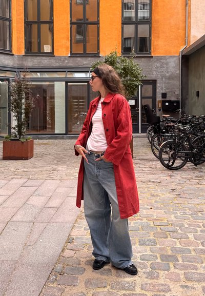 Abrigo largo rojo, camiseta blanca, vaqueros azules sueltos, zapatos negros. Escenario: área de adoquines con bicicletas y edificios modernos de fondo.