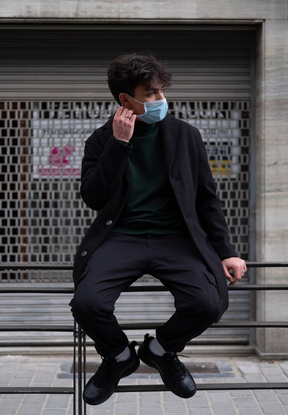 Black overcoat, green turtleneck, black trousers, and sneakers. A light blue mask is worn while seated on a metal railing. Urban background featuring graffiti.