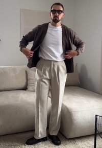 Homme portant des lunettes, un pantalon beige, un t-shirt blanc et un cardigan marron, ajustant son cardigan dans un salon aux tons neutres.