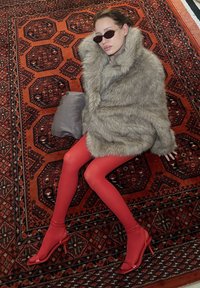 Femme allongée sur un tapis à motifs, portant un manteau en fausse fourrure gris surdimensionné, des collants rouges, des talons rouges et des lunettes de soleil foncées, avec un sac à main gris à proximité.