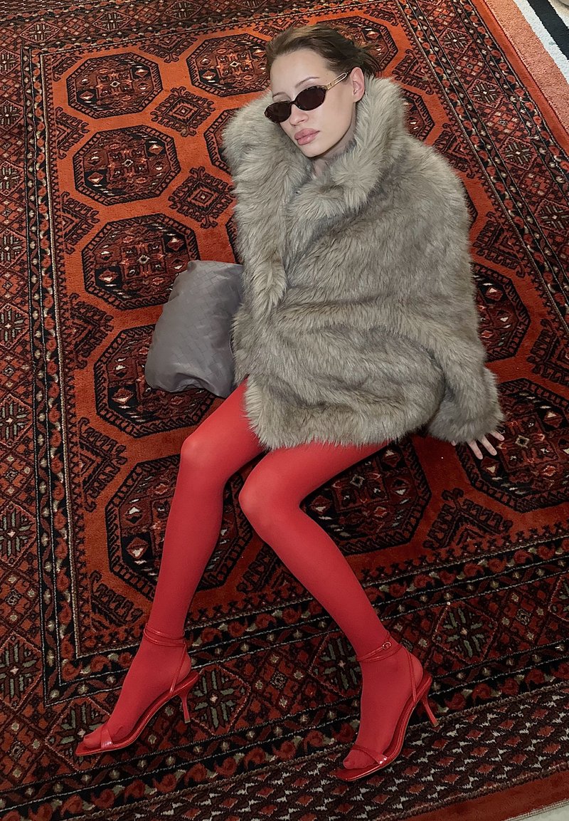 Femme allongée sur un tapis à motifs, portant un manteau en fausse fourrure gris surdimensionné, des collants rouges, des talons rouges et des lunettes de soleil foncées, avec un sac à main gris à proximité.