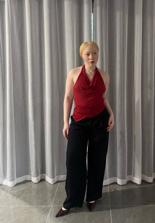 Oseba s svetlimi lasmi, oblečena v rdečo zgornjo majico na vrvico, črne široke hlače in svetleče čevlje, stoji ob sivih zavesah.