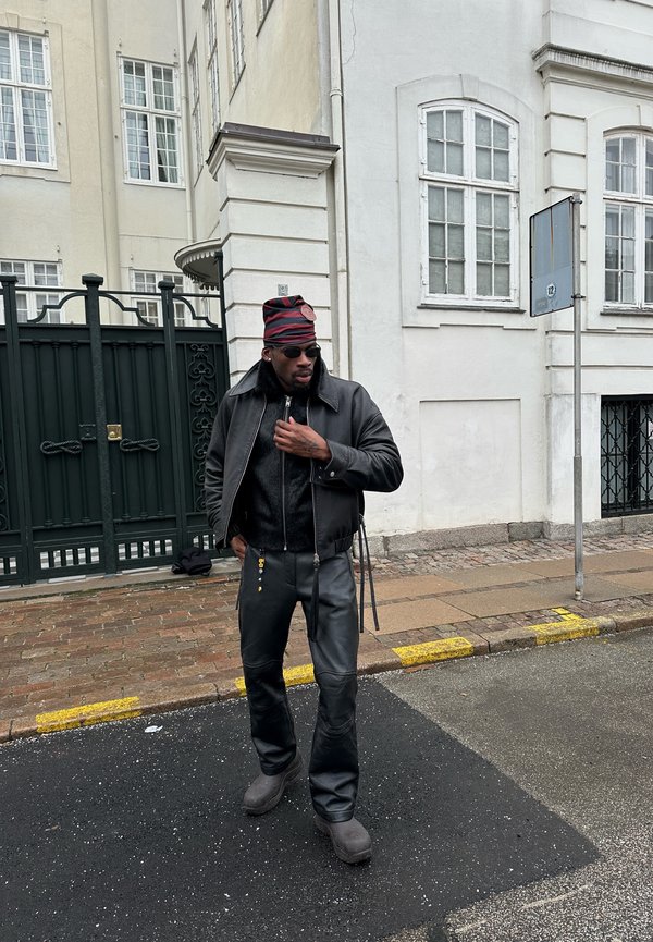 Homme portant une veste en cuir noire, un pantalon, des bottes, des lunettes de soleil et un chapeau à rayures rouges, debout dans une rue près d'un bâtiment blanc et d'une grille.