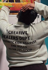 Sweatshirt verde claro com texto em preto em destaque nas costas, escrito "CREATIVE DEALERS DEPT. AESTHETIC KIDS AGAINST THE RULES SO FAR / SO GOOD".