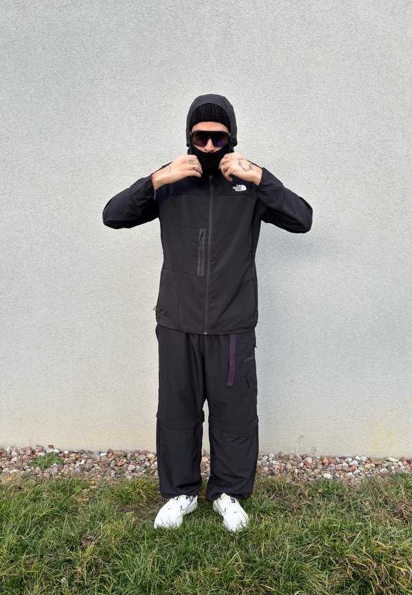 Czarna bluza z zamkiem i kapturem, w zestawie z luźnymi czarnymi spodniami. Cechy obejmują przednią kieszeń i kontrastowy fioletowy pasek. Nosząc okulary przeciwsłoneczne.