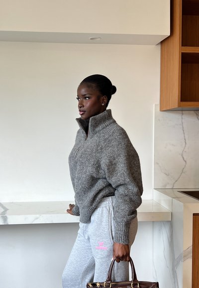 Mujer de piel oscura con suéter gris y pantalones deportivos, de pie en el interior sosteniendo un bolso marrón junto a una encimera de mármol.