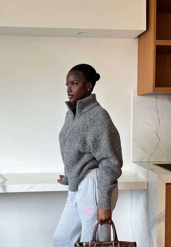 Femme à la peau foncée portant un pull gris et un pantalon de survêtement, debout à l'intérieur tenant un sac à main marron à côté d'un comptoir en marbre.