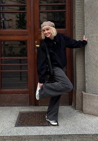 Svart kort jacka, grå vida byxor och silverfärgade sneakers. Linjärt mönstrad beige keps. Står vid en träddörr med ett metallgaller.