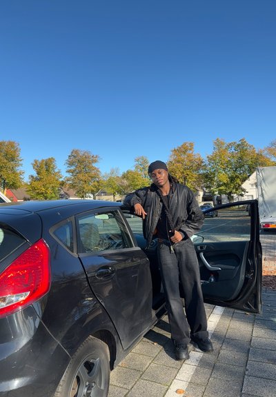 Coche hatchback negro con una puerta abierta. La persona lleva una chaqueta bomber negra y pantalones negros holgados, de pie junto al vehículo.