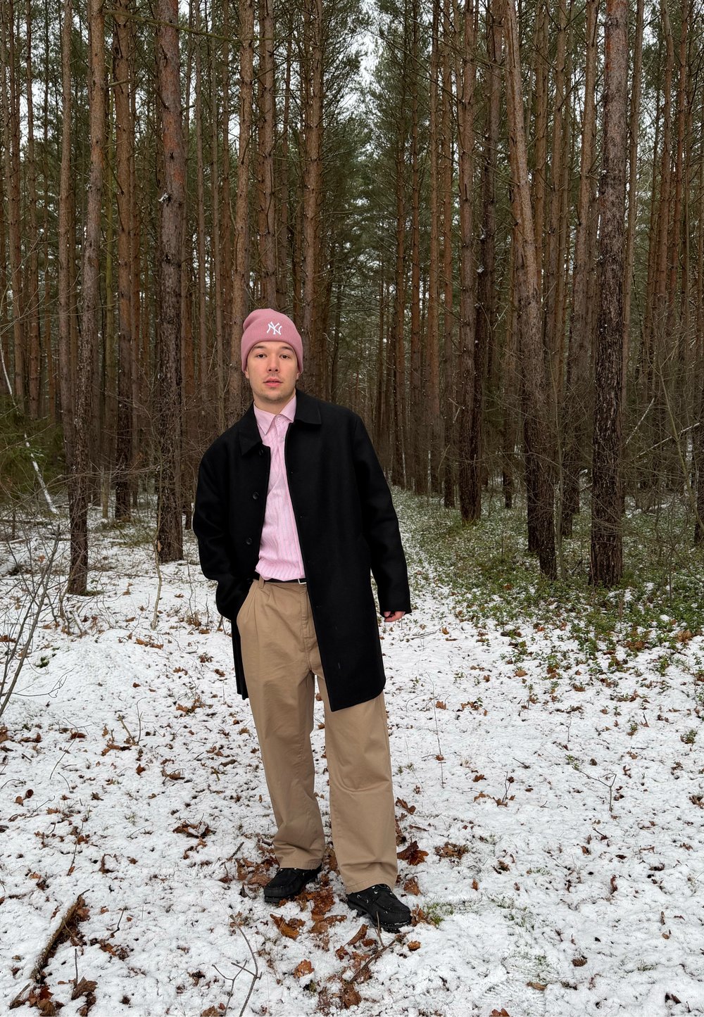 Jeune personne portant un bonnet rose, un manteau noir, une chemise rose et un pantalon beige, debout sur un sol enneigé dans une forêt dense de pins.