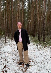 Jeune personne portant un bonnet rose, un manteau noir, une chemise rose et un pantalon beige, debout sur un sol enneigé dans une forêt dense de pins.