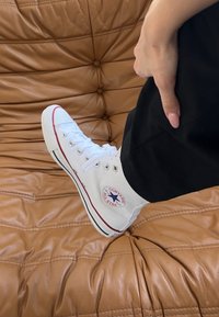 White high-top Converse sneakers with red accents and silver eyelets, featuring a circular logo patch on the outer side.