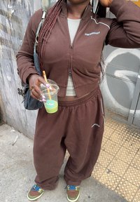 Person wearing a brown tracksuit and colourful sneakers, holding a green drink in a clear cup with a straw, standing on a tiled pavement.