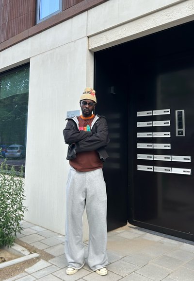 Una persona con un sudadera oversized gris claro y pantalones de chándal, una camiseta marrón y un gorro beis está de pie junto a una entrada oscura con buzones.