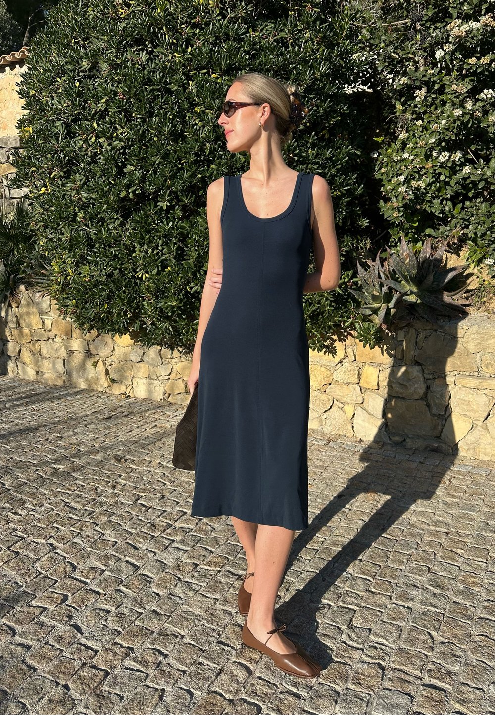Vestido azul marinho sem mangas, com corte até ao joelho, textura suave e decote redondo, combinado com sapatos planos castanhos e uma bolsa texturizada.