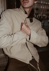 Bēšs viegls džemperis ar rāvējslēdzēja aizdari, ar kordurojas apkakli un redzamu šuvju apdari. Valkāts pāri brūnai drēbju daļai, rokas noregulē rāvējslēdzēju.