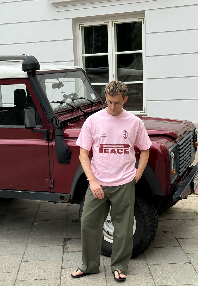 Homme en T-shirt rose et pantalon vert se tient la tête baissée à côté d'un véhicule tout-terrain bordeaux dans une rue de la ville.