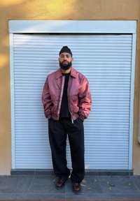 Moški v roza satenasti bomber jakni, črni majici in širokih črnih hlačah s črtami stoji pred belimi rolo vrati.