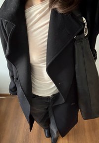 Femme portant un manteau noir, un haut crème froncé, un pantalon noir, des chaussures noires, et portant un sac à bandoulière en cuir noir, debout sur un sol en bois.