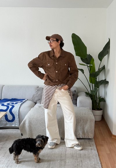 Persona con chaqueta marrón, pantalones blancos y gorra, de pie junto a un perro pequeño negro y marrón en una sala de estar con sofá gris y planta alta y verde.