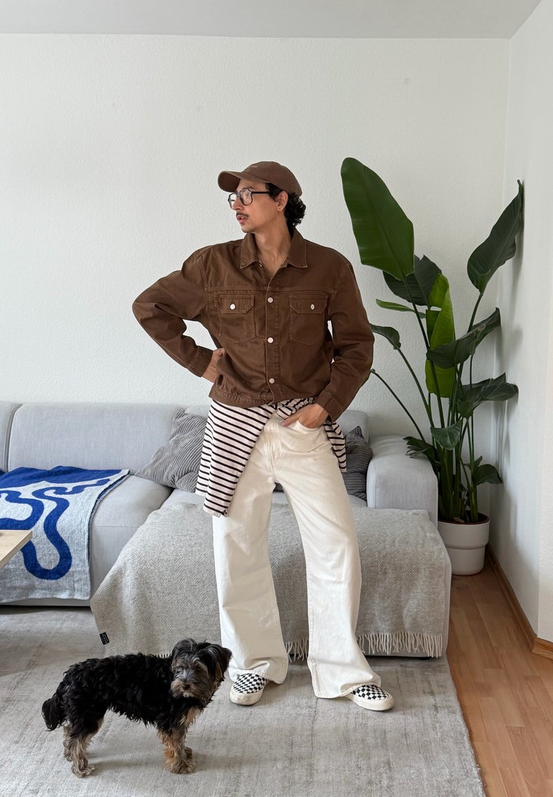 Personne en veste marron, pantalon blanc et casquette se tient à côté d'un petit chien noir et marron dans un salon avec un canapé gris et une grande plante verte.