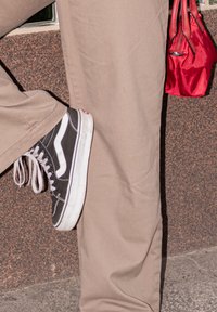 Fekete Vans sportcipő fehér részletekkel, bézs nadrággal viselve, texturált felülettel. Mellette részben látható egy piros kézitáska.