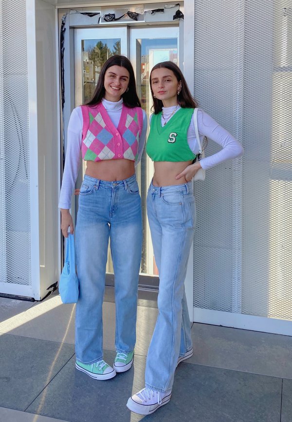 Dos mujeres están de pie una al lado de la otra. Una lleva un chaleco a rayas en tonos rosa y verde, la otra una camiseta corta verde con una "S". Ambas visten jeans de mezclilla claros y zapatillas.