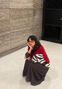 Robe marron texturée avec un pull rayé marron et blanc, châle rouge sur les épaules, et lunettes de soleil jaunes. Cadre intérieur avec des murs gris.