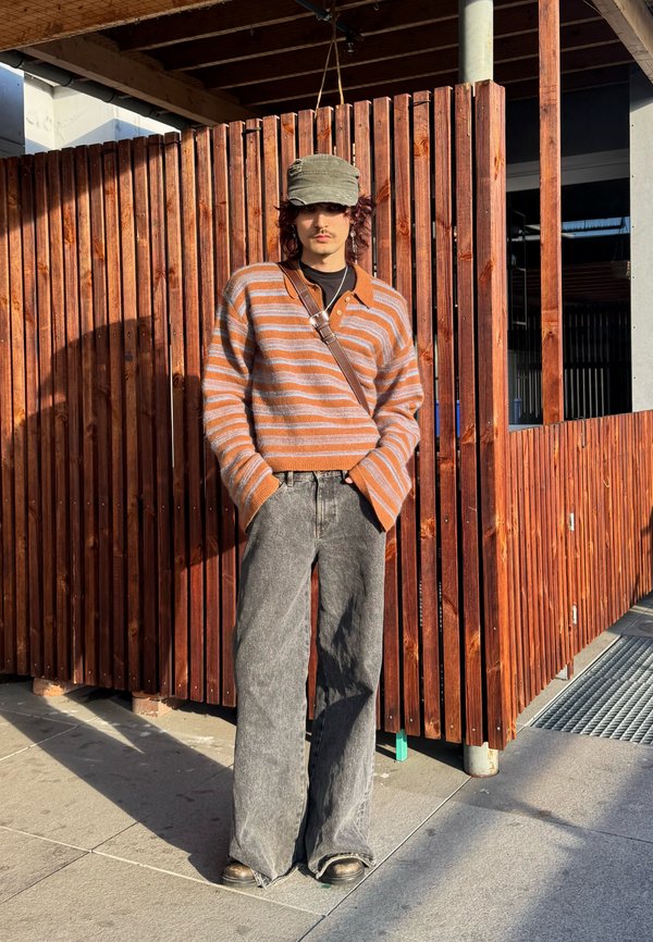 Person trägt einen gestreiften braun-grauen Pullover, graue Jeans mit weitem Bein, grüne Mütze und eine Umhängetasche und steht bei Sonnenlicht an einem Holzzaun.