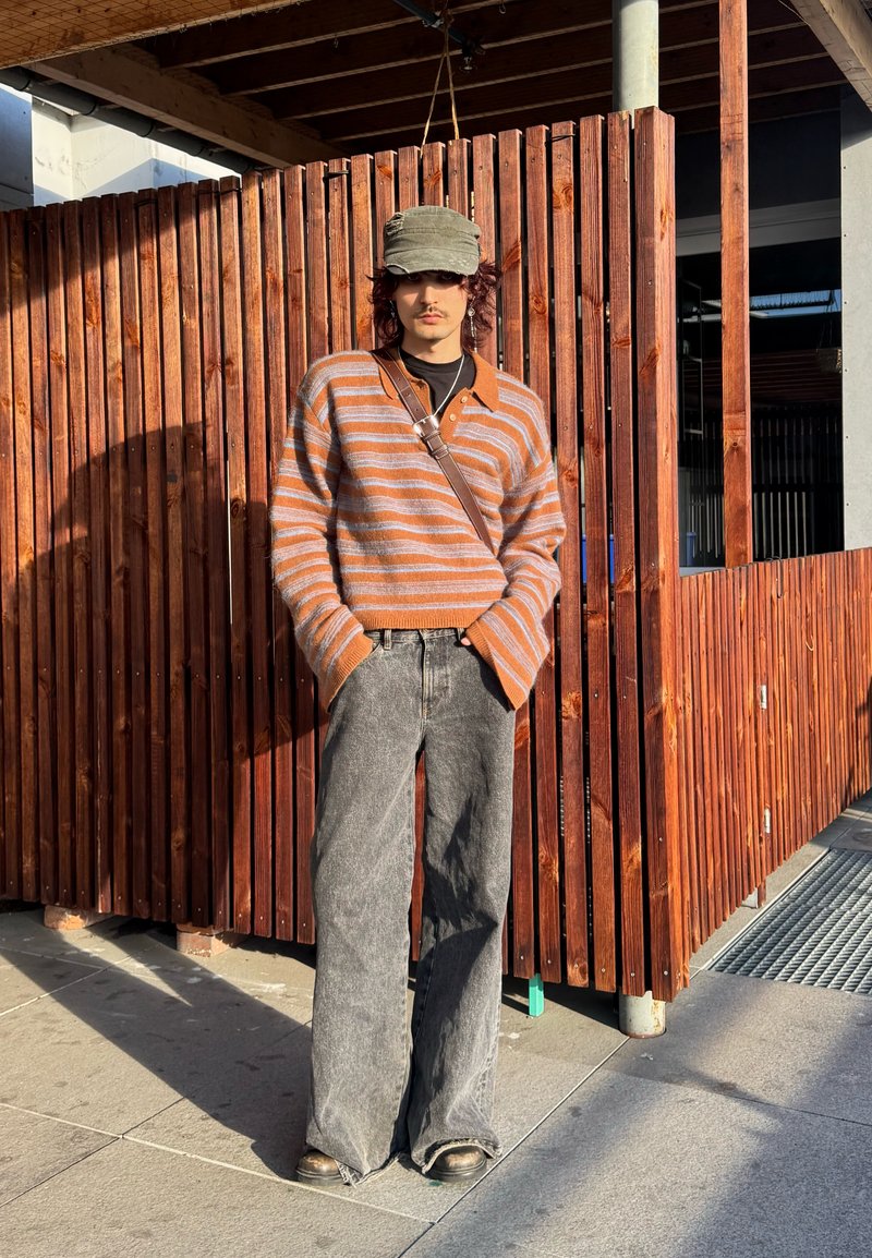 Persona che indossa un maglione a righe marrone e grigio, jeans grigi a gamba larga, berretto verde e borsa a tracolla, in piedi contro una recinzione di legno al sole.