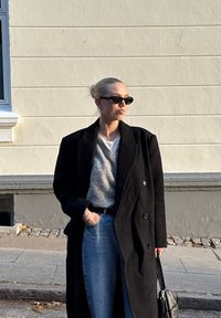 Μαύρο διπλόπετο παλτό πάνω από γκρίζο πουλόβερ, συνδυασμένο με ανοιχτό γαλάζιο τζιν. Αξεσουάρ περιλαμβάνουν μαύρα γυαλιά ηλίου και μια τσάντα.