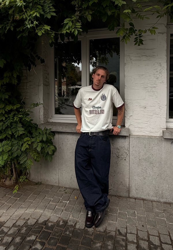 A young man with tattoos leans against a windowsill outside a white building, wearing a white Berlin t-shirt, black baggy trousers, and dark shoes.