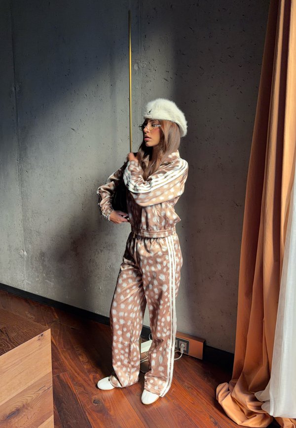 Joven mujer con chándal marrón con manchas, zapatillas blancas y gorro blanco peludo, de pie cerca de una pared gris y cortinas naranjas en el interior.