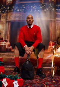 Pull rouge à torsades, short plissé noir, chaussettes rouges, chaussures en cuir noir, tenant un sac noir avec des accents dorés, sur un fond festif.