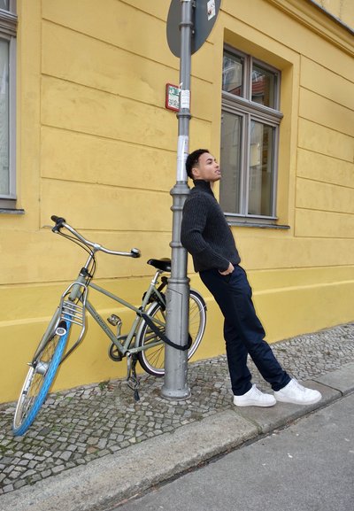 Bicicleta con un marco verde y acentos azules, apoyada contra un poste gris en una calle de adoquines. Una persona está de pie cerca, vestida con ropa oscura.