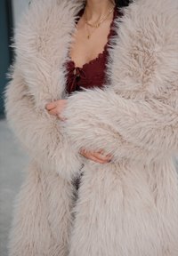 Textúrovaný kabát z umelého kožušiny v svetlej béžovej farbe, s dlhými rukávmi a nadýchaným vzhľadom. Nosí sa cez bordový top s čipkovými akcentmi.