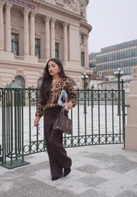 Maglione a maglia con stampa leopardata e dettagli beige, pantaloni a gamba larga marrone scuro, stivaletti neri e borsa marrone. Tiene in mano una rivista.