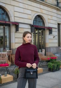 Bordeaux geribbelde gebreide trui met hoge kraag, grijze broek en een kleine zwarte handtas met textuuraccenten. Buitensetting met meubels.