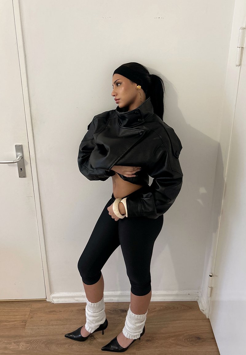 Femme portant une veste en cuir noire courte, des leggings noirs, des guêtres blanches et des talons noirs, debout contre un mur blanc à l'intérieur.