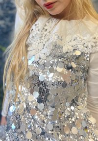 Robe en sequins argentés avec une surface texturée, présentant un col en dentelle et des motifs floraux. Gros plan sur des sequins circulaires brillants et un tissu blanc.