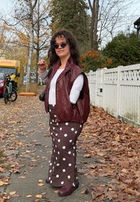 Vīna krāsas ādas veste pār baltu topu, kombinēta ar brūnu svārku ar punktiņiem. Tumši brūni zābaki. Fona attēlā rudens lapas un balts žogs.