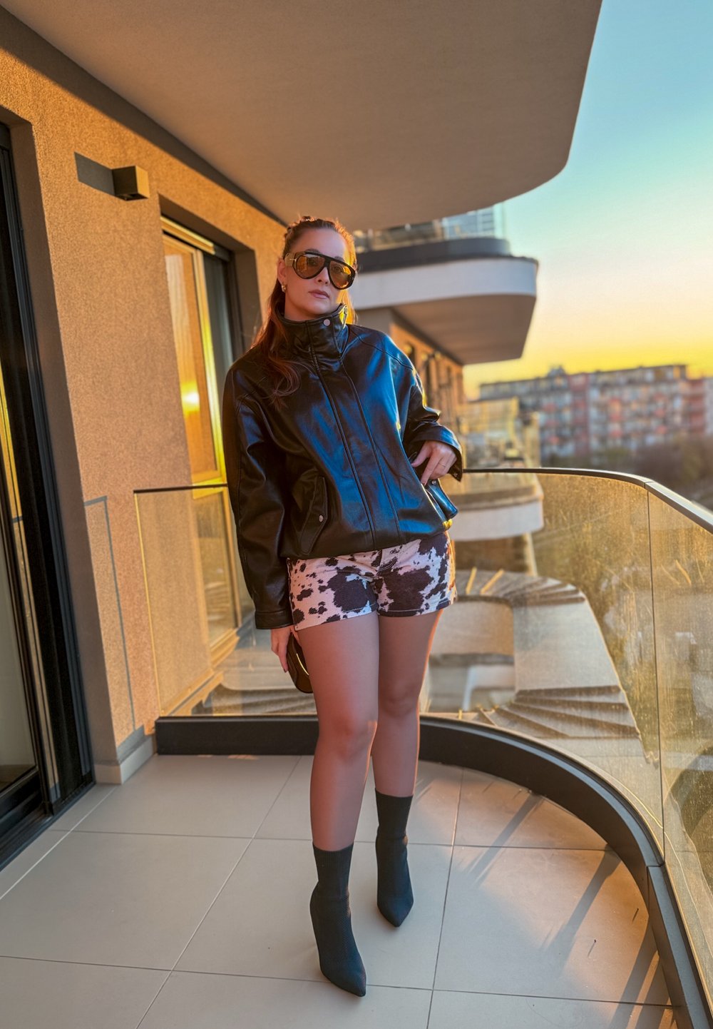 Černá kožená bunda, krátké kalhoty s potiskem kravského vzoru a tmavé kotníkové boty. Styl doplňují sluneční brýle.